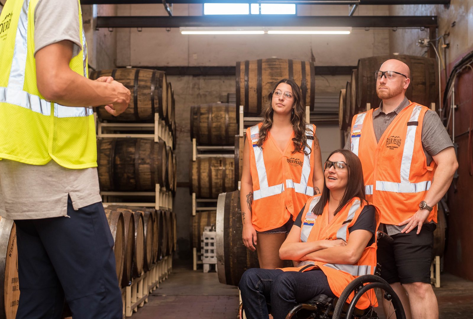 Three workers casually dressed, two women (one standing, left, and one sitting in a wheelchair, center front) and a man, stand in a warehouse full of barrels wearing protective glasses and orange vests. A man with a yellow vest stand off in in front of them. They look to him.