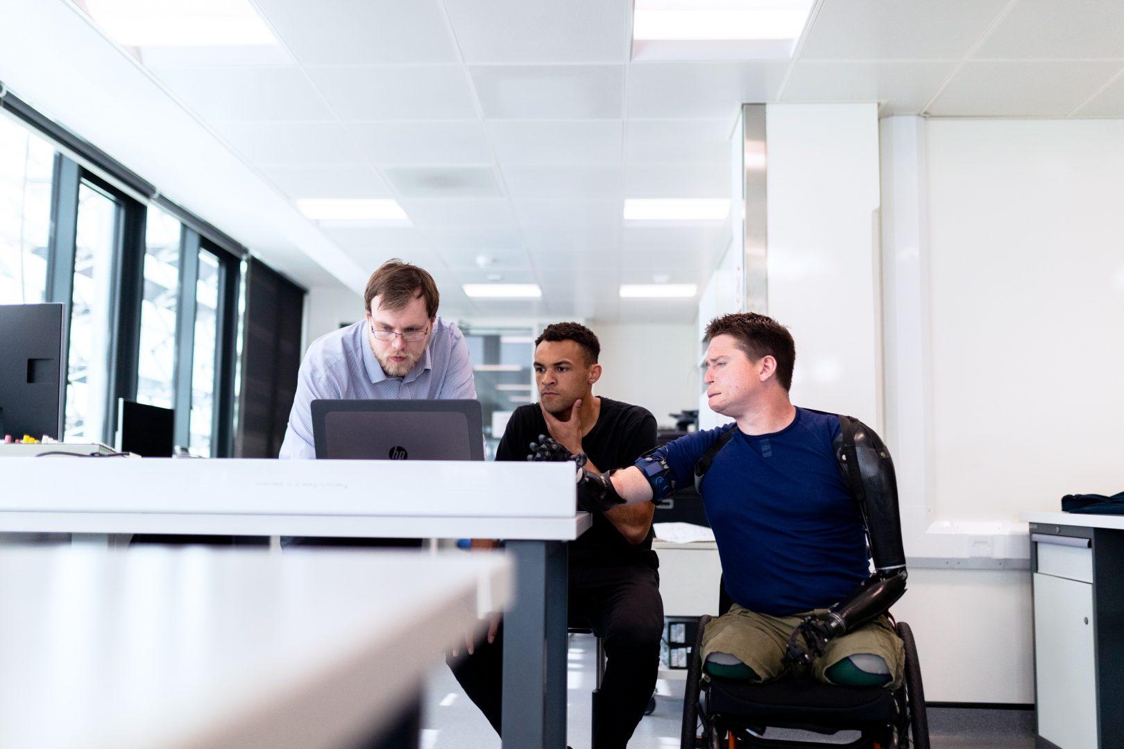 Three men (From left to right: Caucaian male wearing blue button down with brown beard hair and bard leans down over a laptop, staring at the computer. An African American male holding his chin with one hand , sits in a chair and looks at the same laptop. A Caucaian male, sitting in a wheelchair, with two prosthetic arms and legs looks over at the same laptop.) Look at a laptop intensely. The are in a brightly lit white office.