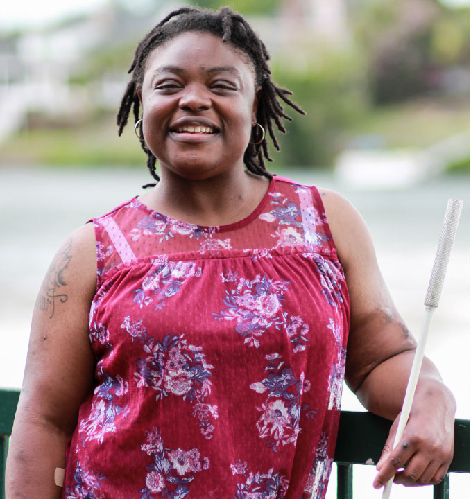 An African American woman, wearing a red floral shirt, holds a cane in her hand as she smiles wide for the camera.