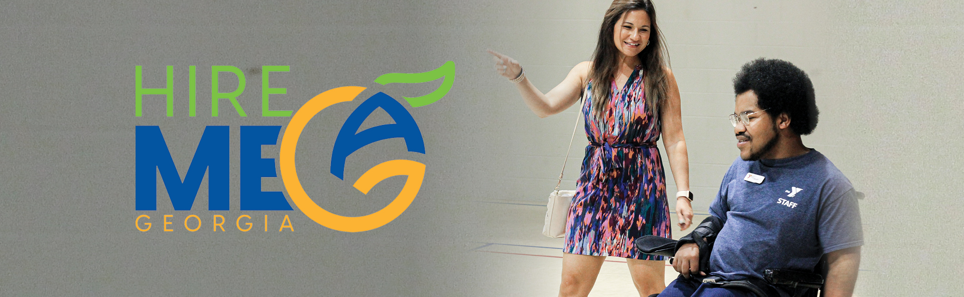 To the left of the image, in grey space sits a green, blue, and orange stylized Stacked Hire Me GA logo. Beside is an image of a caucasian woman, wearing a multicolored dress, pointing to her right as she walks beside an African American male in a wheelchair.