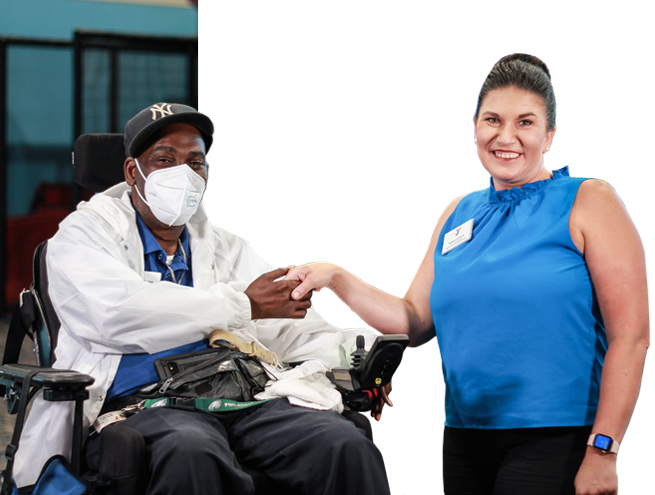 A man and woman shake hands while looking toward a camera. The man, to the left, a middle aged male wearing a NY black cap, a white face mask, blue polo shirt and white jacket, sits in a wheelchair with his hand extended. A caucasian female wearing her hair up in a bun, a blue sleeveless shirt and black pants, extends her hand to meet the mans. She smiles.